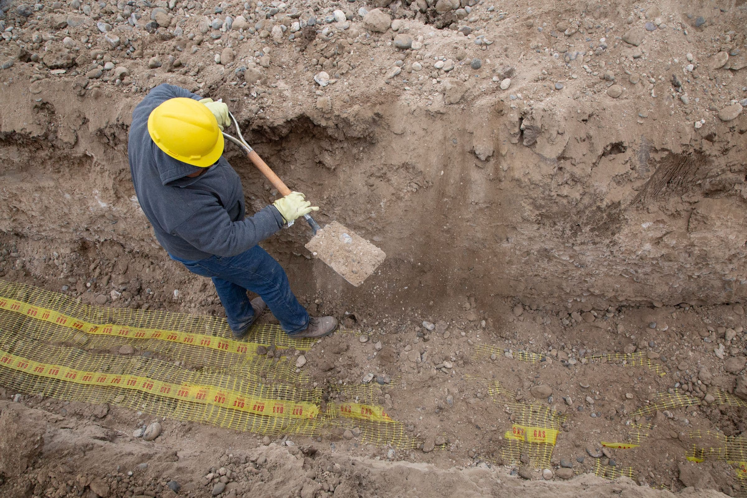 Looking down on a worker in a yellow hard hat standing in a narrow trench, driving a spade into the exposed soil wall, with survey tape running along the ground above.