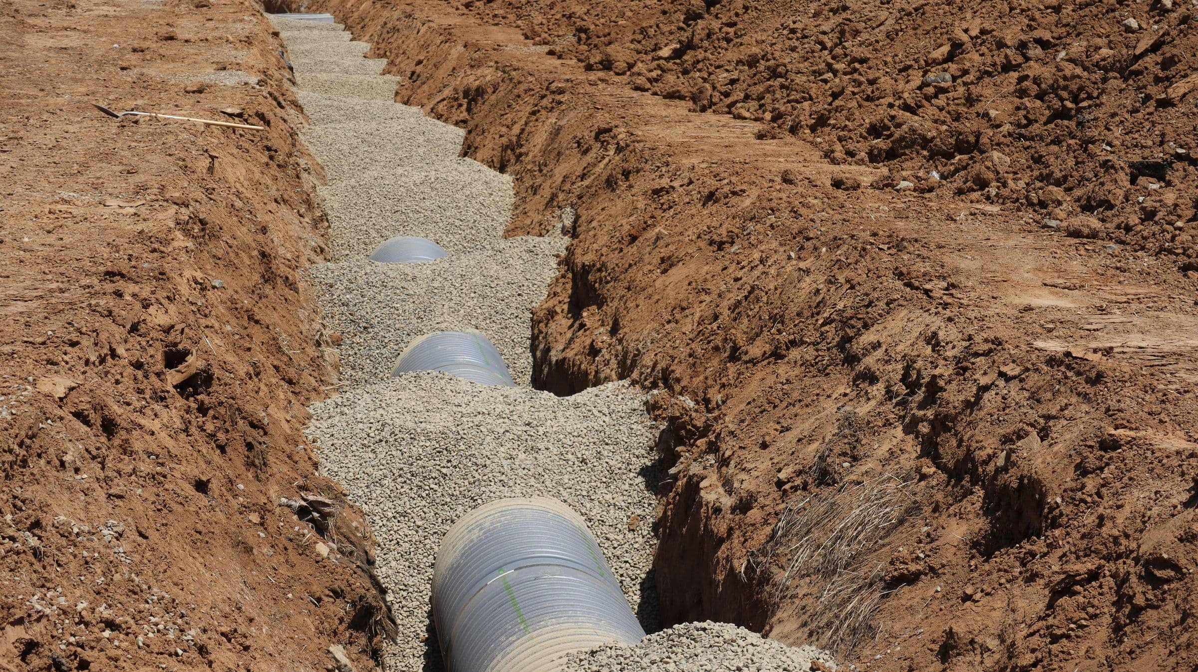 A perforated corrugated drainage pipe laid along the bottom of a trench in reddish clay soil and surrounded by clean pale drainage gravel, running away from the camera.