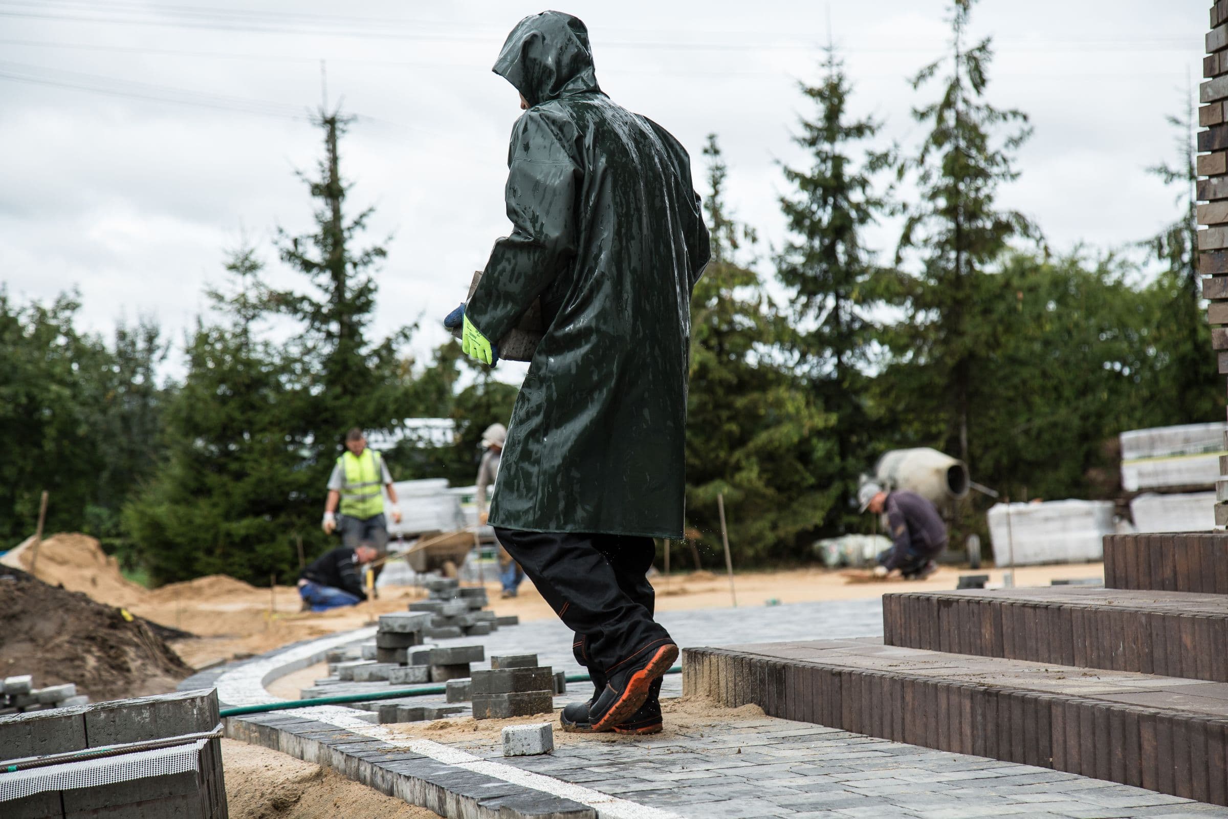 A worker in a full-length dark rain cape walking across a partly finished paved area, with two more of the crew working further back beside a sand pile and a row of young conifers under a grey sky.