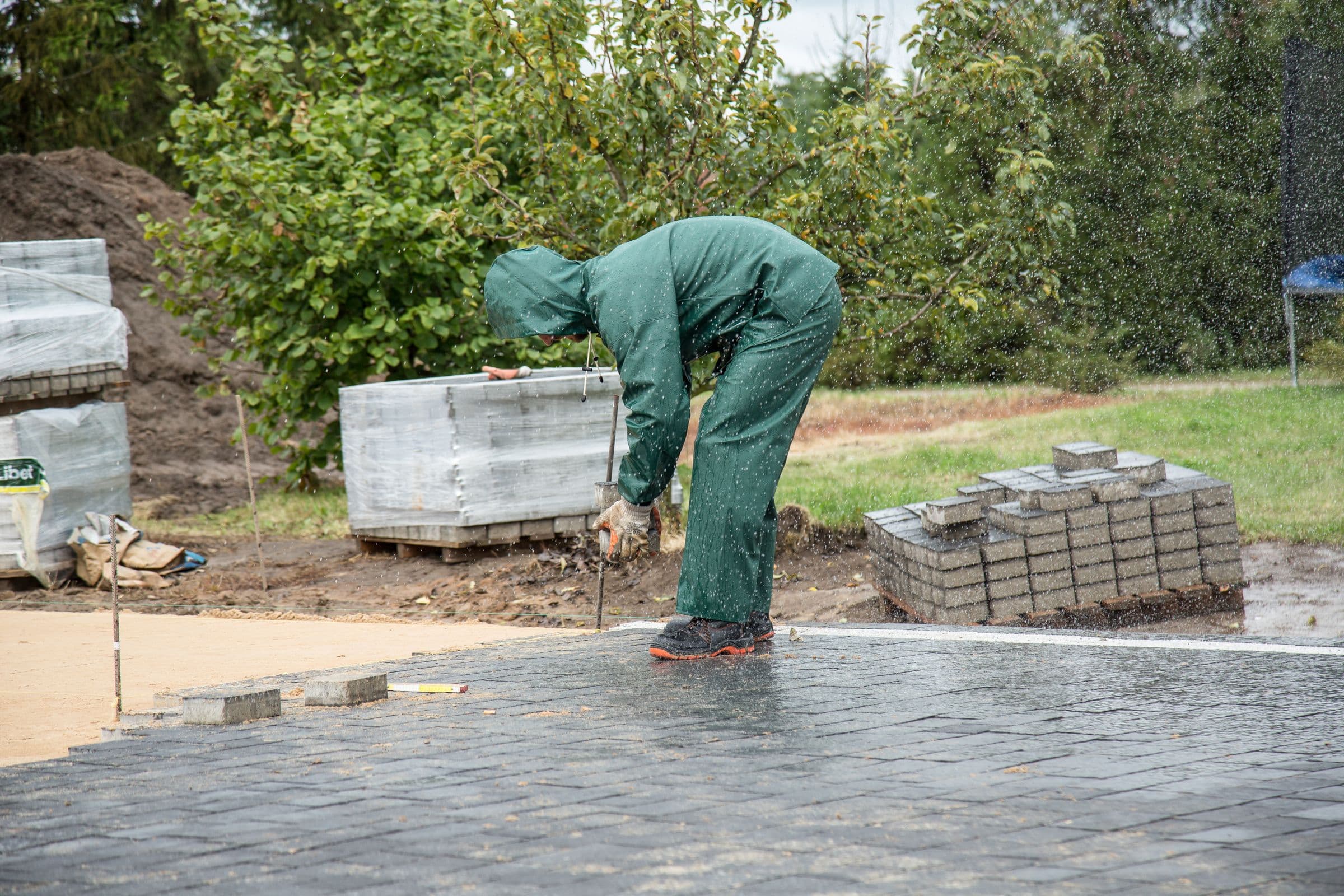 A worker in green waterproof trousers and jacket bent double over a string line on a half-laid block paving area, with rain visibly falling, a screeded sand bed to one side and stacked pallets of pavers behind.