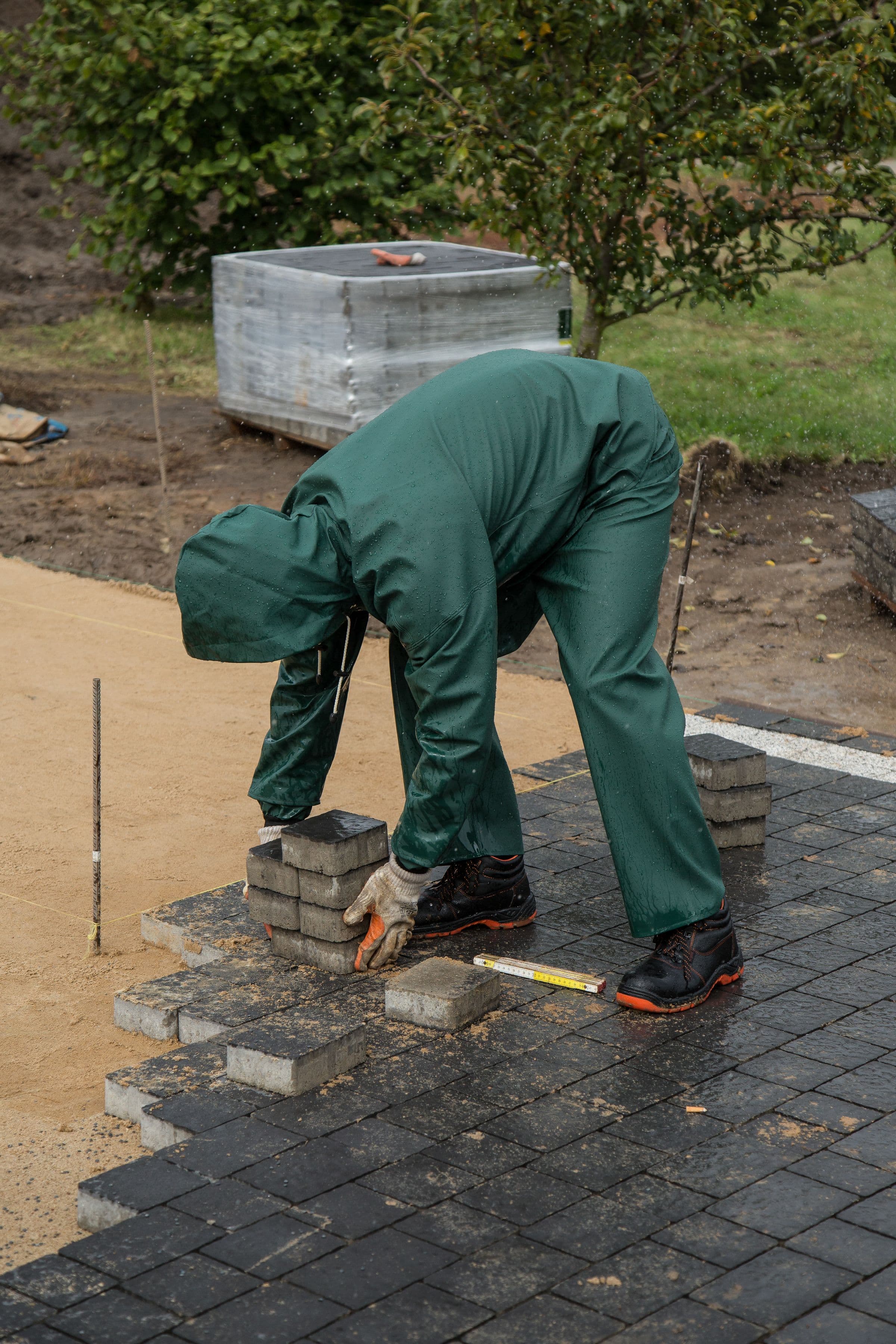 A worker in green waterproofs kneeling to place a paving block against an already-laid course, with a rubber mallet and a stack of blocks beside him on the screeded sand.