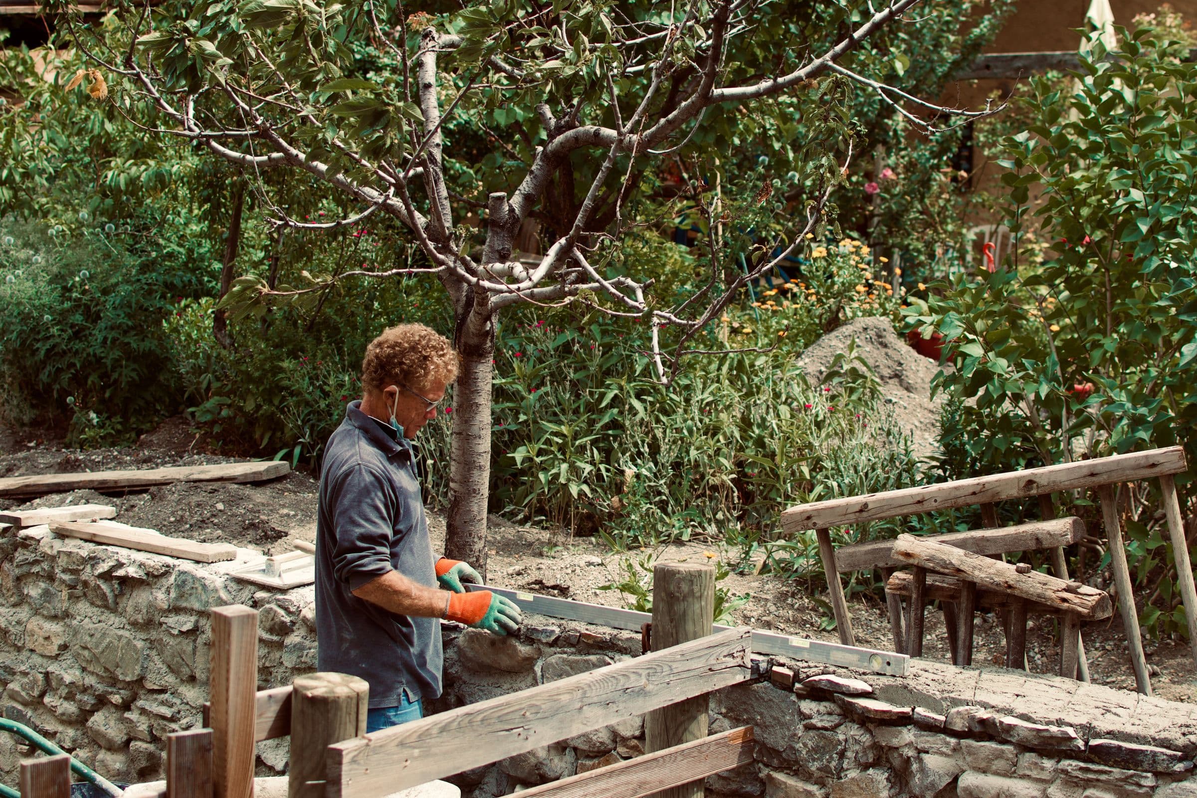 A man in work gloves and a grey shirt building a low stone retaining structure behind timber shuttering in a garden, an old fruit tree and a wheelbarrow behind him, looking down at the stone he is placing.